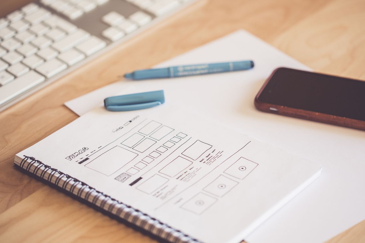 heros-img A neat workspace featuring a sketchpad with wireframes, a smartphone, and a keyboard on a wooden desk.